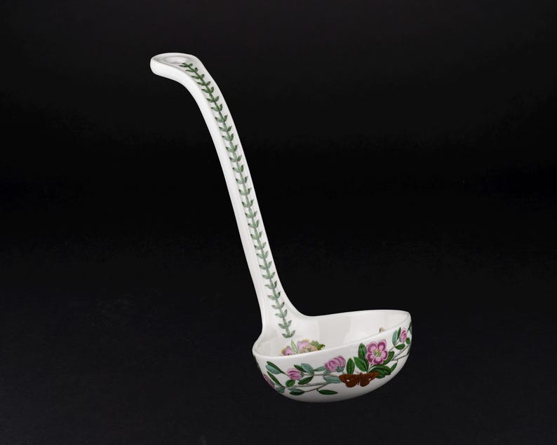 May include: A white ceramic ladle featuring a long handle decorated with a green leaf vine pattern. The bowl of the ladle is adorned with a colorful botanical illustration of pink flowers, green leaves, and a small brown butterfly on the exterior.