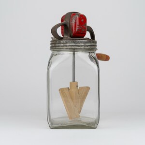 May include: A vintage glass butter churn featuring a square clear glass jar, a metal lid with a red painted top mechanism, and a wooden paddle agitator attached to a central metal rod. A small wooden handle extends from the side of the metal lid.