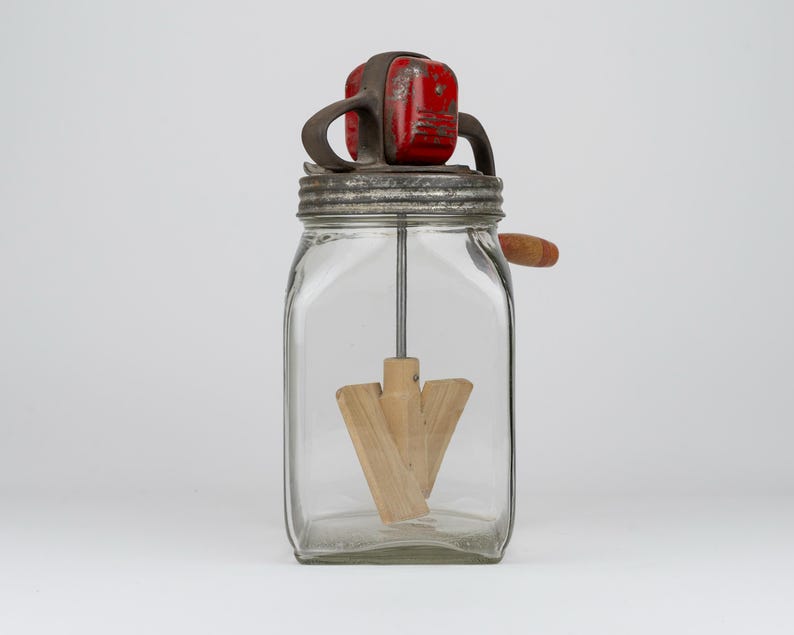 May include: A vintage glass butter churn featuring a square clear glass jar, a metal lid with a red painted top mechanism, and a wooden paddle agitator attached to a central metal rod. A small wooden handle extends from the side of the metal lid.