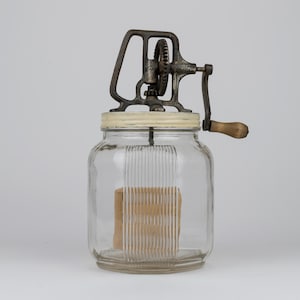 K&ouml;nnte beinhalten: Ein antiker Butterbereiter aus Glas mit einem klaren, gerippten Glasbeh&auml;lter und einem wei&szlig;en Deckel. Der obere Mechanismus besteht aus dunklem, verwittertem Metall mit einem Getriebe und einem Holzgriff an einer Kurbel zur manuellen Bedienung.