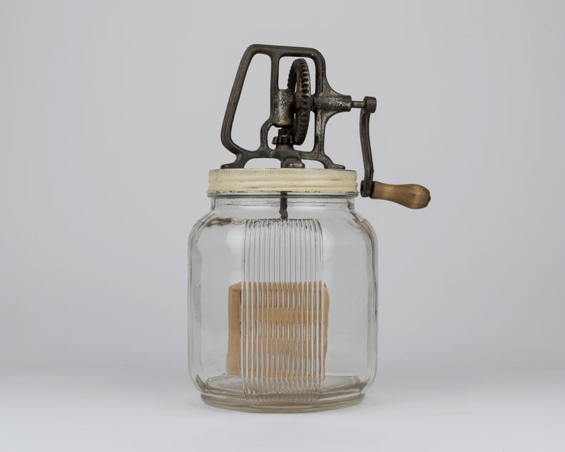 K&ouml;nnte beinhalten: Ein antiker Butterbereiter aus Glas mit einem klaren, gerippten Glasbeh&auml;lter und einem wei&szlig;en Deckel. Der obere Mechanismus besteht aus dunklem, verwittertem Metall mit einem Getriebe und einem Holzgriff an einer Kurbel zur manuellen Bedienung.