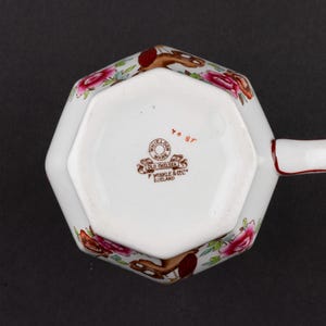 K&ouml;nnte beinhalten: Eine wei&szlig;e, achteckige Keramiktasse mit Henkel, verziert mit einem Blumenmuster aus rosa Rosen und braunen Akzenten. Der Boden zeigt einen Stempel mit "Whieldon Ware Old Chelsea F Winkle & Co Ltd England" und den handgeschriebenen roten Text "70 87".