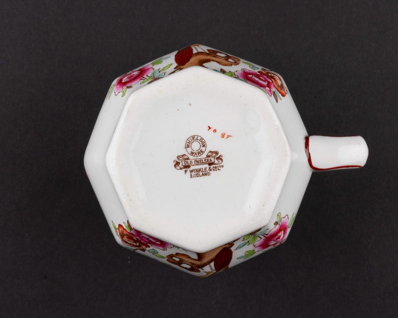 K&ouml;nnte beinhalten: Eine wei&szlig;e, achteckige Keramiktasse mit Henkel, verziert mit einem Blumenmuster aus rosa Rosen und braunen Akzenten. Der Boden zeigt einen Stempel mit "Whieldon Ware Old Chelsea F Winkle & Co Ltd England" und den handgeschriebenen roten Text "70 87".