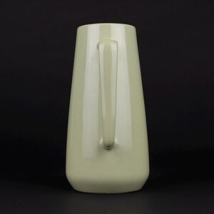 May include: A pale sage green ceramic pitcher with a smooth, glossy finish. The vessel features a tapered, conical body shape that widens towards the base and includes a single vertical handle attached to the side for gripping and pouring.