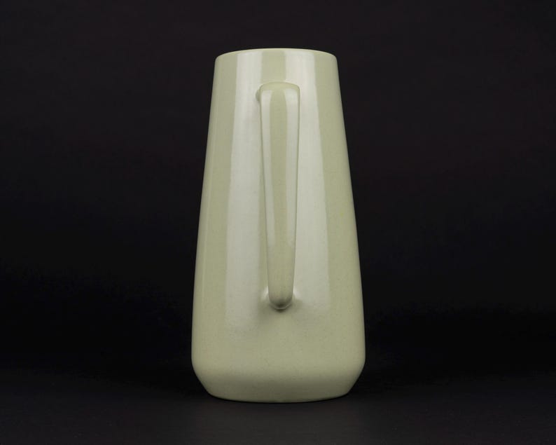 May include: A pale sage green ceramic pitcher with a smooth, glossy finish. The vessel features a tapered, conical body shape that widens towards the base and includes a single vertical handle attached to the side for gripping and pouring.