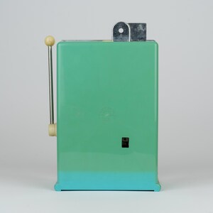 May include: A rectangular mint green kitchen appliance with a vertical metal lever on the left side, featuring a cream-colored knob at the top. The device has a small black square switch on the front and a silver metal mechanism protruding from the top.