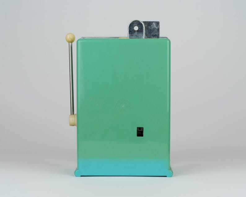 May include: A rectangular mint green kitchen appliance with a vertical metal lever on the left side, featuring a cream-colored knob at the top. The device has a small black square switch on the front and a silver metal mechanism protruding from the top.
