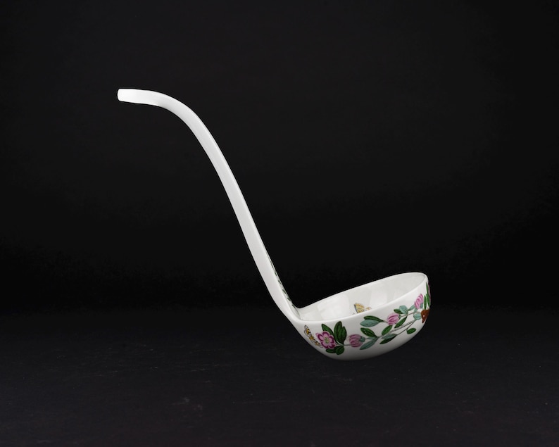 May include: A white ceramic soup ladle featuring a delicate hand-painted floral pattern with pink blossoms, green leaves, and small golden butterfly accents. The ladle has a long, curved handle and a deep, rounded bowl for serving liquids.