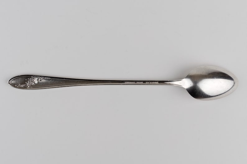 May include: A silver-colored metal spoon with a long, slender handle featuring an embossed floral pattern at the end. The handle is engraved with the text "SILVERPLATE" and "WM ROGERS MFG CO" in small, clear capital letters near the bowl.