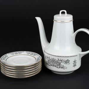 K&ouml;nnte beinhalten: Eine wei&szlig;e Kaffeekanne aus Porzellan mit Deckel und ein dazu passender Stapel aus sechs Untertassen. Das Set zeigt ein schwarzes Blumenmuster am Kannenboden und an den R&auml;ndern der Untertassen, akzentuiert durch einen d&uuml;nnen Goldrand.