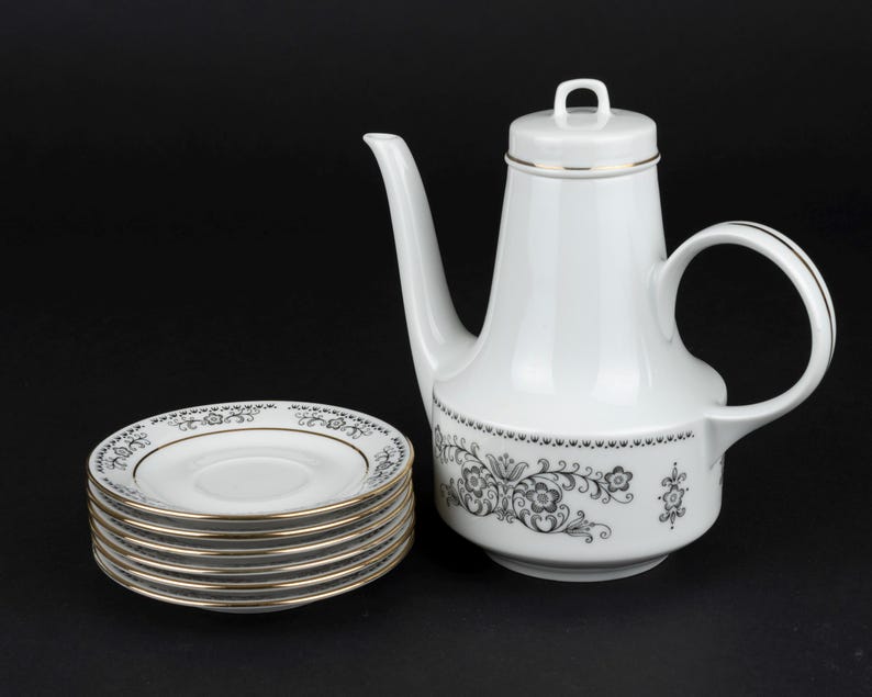 K&ouml;nnte beinhalten: Eine wei&szlig;e Kaffeekanne aus Porzellan mit Deckel und ein dazu passender Stapel aus sechs Untertassen. Das Set zeigt ein schwarzes Blumenmuster am Kannenboden und an den R&auml;ndern der Untertassen, akzentuiert durch einen d&uuml;nnen Goldrand.