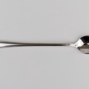 May include: A silver-toned metal spoon featuring an elongated handle with a decorative embossed floral pattern at the tip and a smooth, oval-shaped bowl. The metallic surface has a polished finish with subtle reflective qualities throughout the piece.