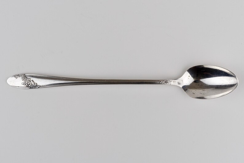 May include: A silver-toned metal spoon featuring an elongated handle with a decorative embossed floral pattern at the tip and a smooth, oval-shaped bowl. The metallic surface has a polished finish with subtle reflective qualities throughout the piece.