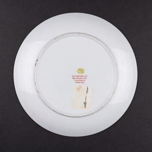 May include: The underside of a white ceramic plate featuring a circular gold sticker and red printed text that reads: NOT FOR FOOD USE. MAY POISON FOOD. FOR DECORATIVE PURPOSES ONLY. A rectangular white adhesive label is also attached to the center.