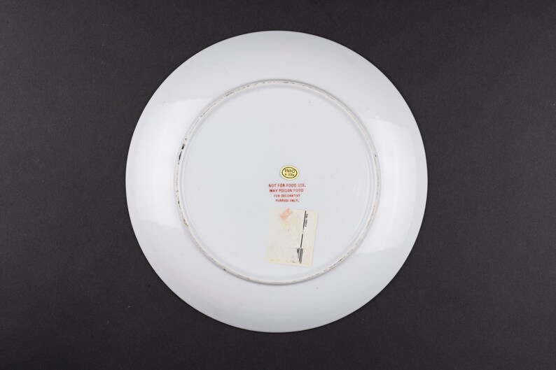 May include: The underside of a white ceramic plate featuring a circular gold sticker and red printed text that reads: NOT FOR FOOD USE. MAY POISON FOOD. FOR DECORATIVE PURPOSES ONLY. A rectangular white adhesive label is also attached to the center.