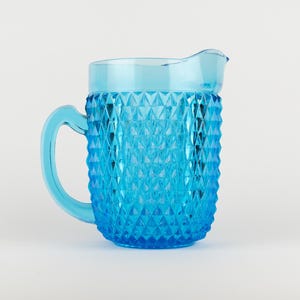 May include: A bright blue glass pitcher featuring a textured diamond pattern across its exterior body. The vessel includes a smooth, curved handle on the left side and a molded pouring spout at the top rim, showcasing a translucent finish.