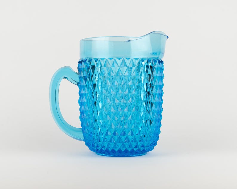 May include: A bright blue glass pitcher featuring a textured diamond pattern across its exterior body. The vessel includes a smooth, curved handle on the left side and a molded pouring spout at the top rim, showcasing a translucent finish.