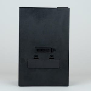 May include: A rectangular black plastic electronic device housing featuring a central battery compartment with a removable cover and a smaller rectangular recessed slot positioned directly above the battery area on the flat surface.