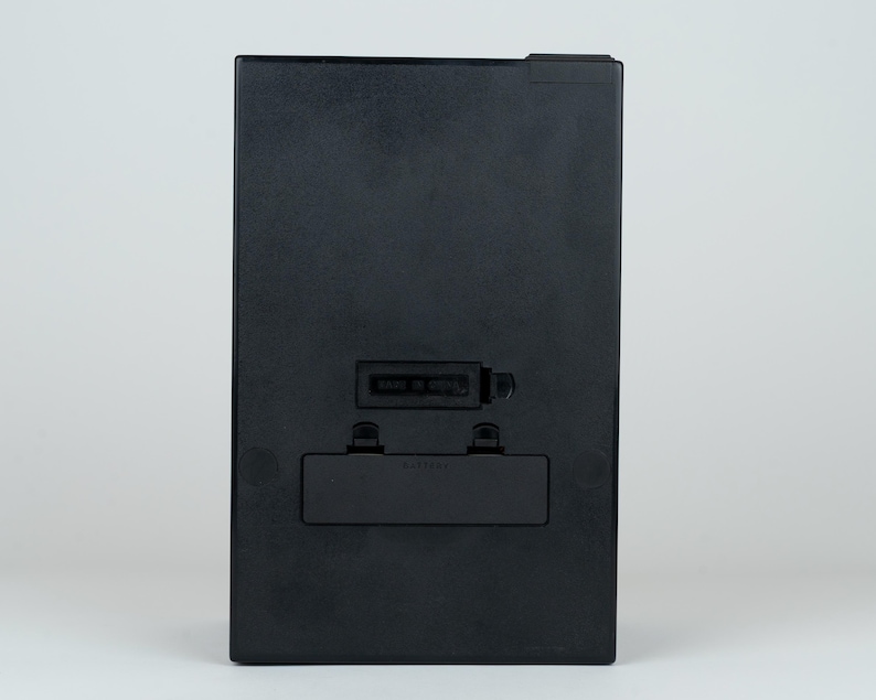 May include: A rectangular black plastic electronic device housing featuring a central battery compartment with a removable cover and a smaller rectangular recessed slot positioned directly above the battery area on the flat surface.