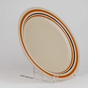 May include: A round, cream-colored ceramic plate featuring a series of concentric circular bands in shades of burnt orange and dark brown. The plate is positioned at an angle, displaying its smooth surface and the distinct color-blocked rim design.