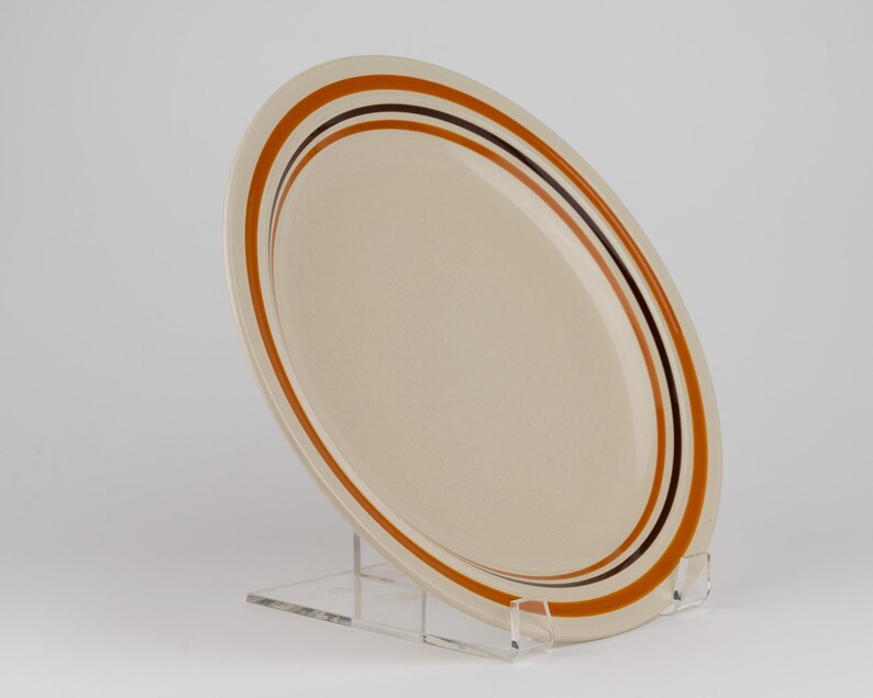 May include: A round, cream-colored ceramic plate featuring a series of concentric circular bands in shades of burnt orange and dark brown. The plate is positioned at an angle, displaying its smooth surface and the distinct color-blocked rim design.