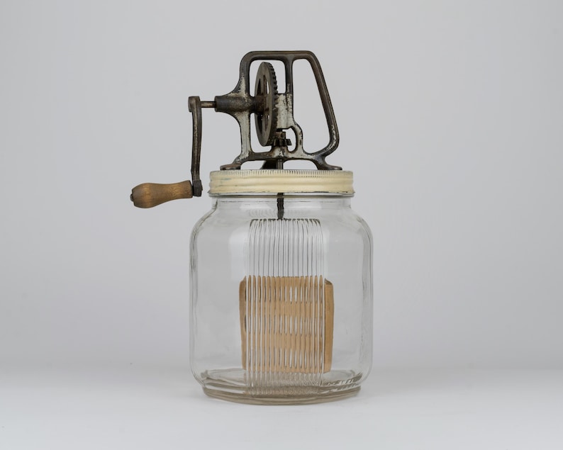 K&ouml;nnte beinhalten: Ein antiker Butterbereiter aus Glas mit einem klaren Glasbeh&auml;lter mit vertikalen Rillen, einem cremefarbenen Metalldeckel und einem dunklen Metall-Kurbelmechanismus mit Holzgriff. Das innere R&uuml;hrwerk ist durch die Glasw&auml;nde deutlich zu erkennen.
