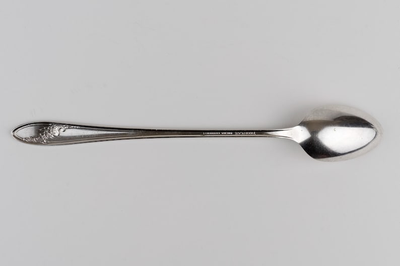 May include: A silver-colored metal spoon featuring an elongated handle with a decorative floral relief pattern at the tip. The handle includes the engraved text "WM. ROGERS MFG. CO. IS" and the bowl of the spoon is smooth and oval-shaped.