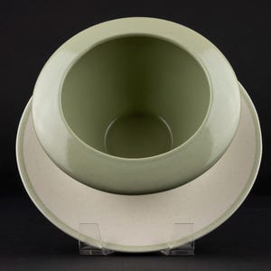 Peut inclure: Un bol en c&eacute;ramique vert sauge avec un int&eacute;rieur lisse et arrondi et un large bord plat. La base ext&eacute;rieure du bol pr&eacute;sente une finition blanc cass&eacute; mouchet&eacute;e. Le bol est plac&eacute; sur un support en acrylique transparent, r&eacute;v&eacute;lant sa forme circulaire.