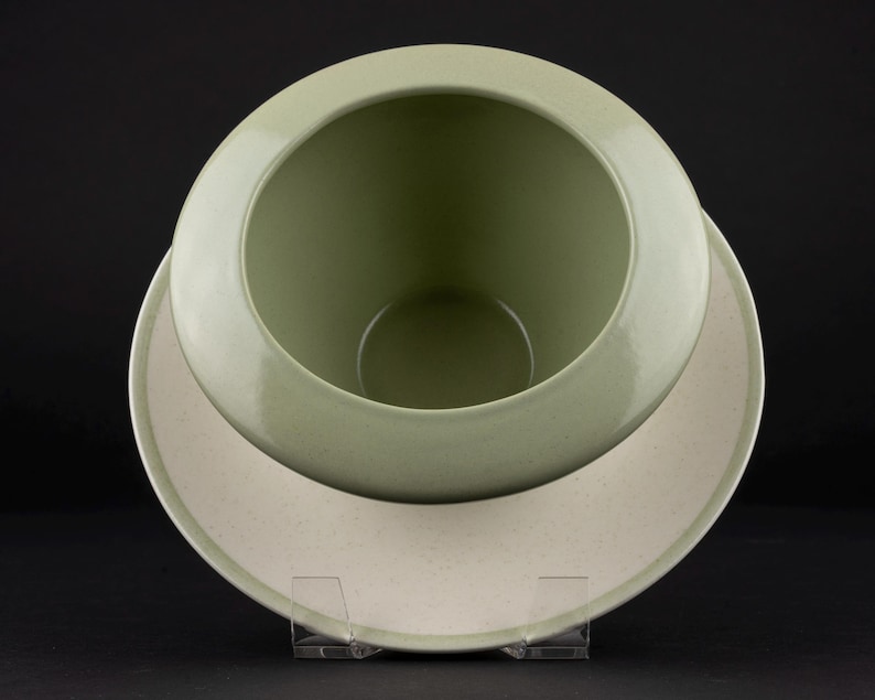 Peut inclure: Un bol en c&eacute;ramique vert sauge avec un int&eacute;rieur lisse et arrondi et un large bord plat. La base ext&eacute;rieure du bol pr&eacute;sente une finition blanc cass&eacute; mouchet&eacute;e. Le bol est plac&eacute; sur un support en acrylique transparent, r&eacute;v&eacute;lant sa forme circulaire.