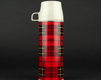 Könnte beinhalten: Eine zylindrische Thermos-Isolierflasche mit einem rot-schwarz-gelben Schottenkaro-Muster. Der Behälter hat einen cremefarbenen Kunststoff-Schraubdeckel, der als Tasse mit einem kleinen Henkel dient. Das Thermos-Logo ist auf der Vorderseite aufgedruckt.