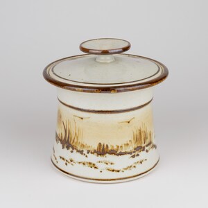 May include: A ceramic canister with a lid featuring a cream-colored base and a brown rim. The body is decorated with a hand-painted landscape scene in shades of brown, depicting tall grasses and birds in flight against a light tan background.