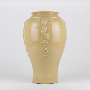 Op de afbeelding: Een glanzende, lichtbeige keramische vaas met een brede, ronde buik en een smalle hals. Het oppervlak is voorzien van verhoogde, gegoten reli&euml;fdecoraties van trapsgewijze bladeren en kleine bloemen met vijf bloemblaadjes.