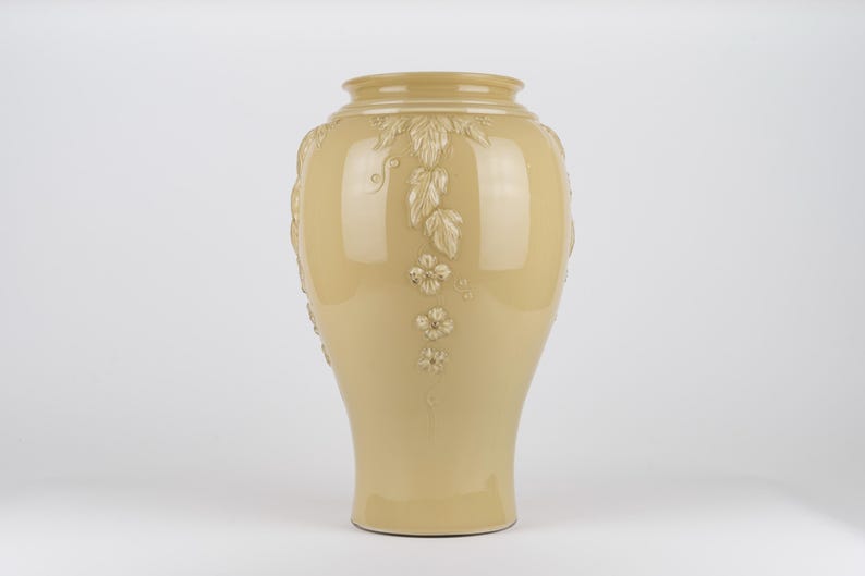 Op de afbeelding: Een glanzende, lichtbeige keramische vaas met een brede, ronde buik en een smalle hals. Het oppervlak is voorzien van verhoogde, gegoten reli&euml;fdecoraties van trapsgewijze bladeren en kleine bloemen met vijf bloemblaadjes.