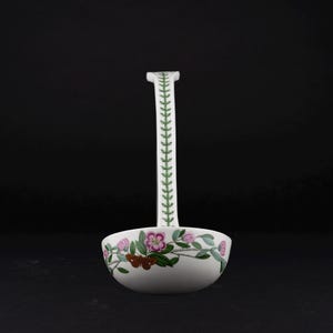 May include: A white ceramic ladle featuring a long handle decorated with a green leaf pattern. The bowl of the ladle is adorned with a colorful botanical illustration of pink flowers, green leaves, and a brown moth against a white background.
