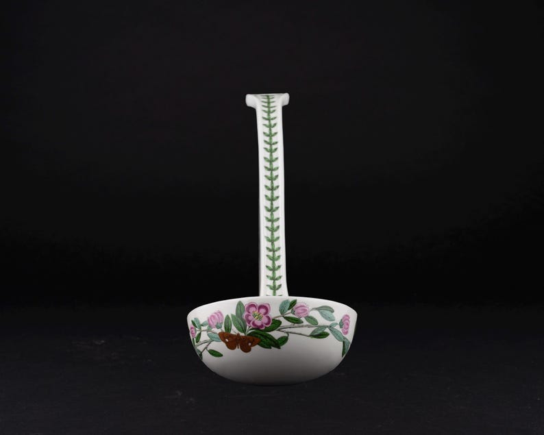 May include: A white ceramic ladle featuring a long handle decorated with a green leaf pattern. The bowl of the ladle is adorned with a colorful botanical illustration of pink flowers, green leaves, and a brown moth against a white background.