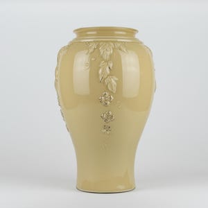Op de afbeelding: Een glanzende, lichtbeige keramische vaas met een brede, ronde buik en een smalle hals. De buitenkant is voorzien van verhoogde, gegoten reli&euml;fdetails van trapsgewijze bladeren en kleine bloemen met vier bloemblaadjes.