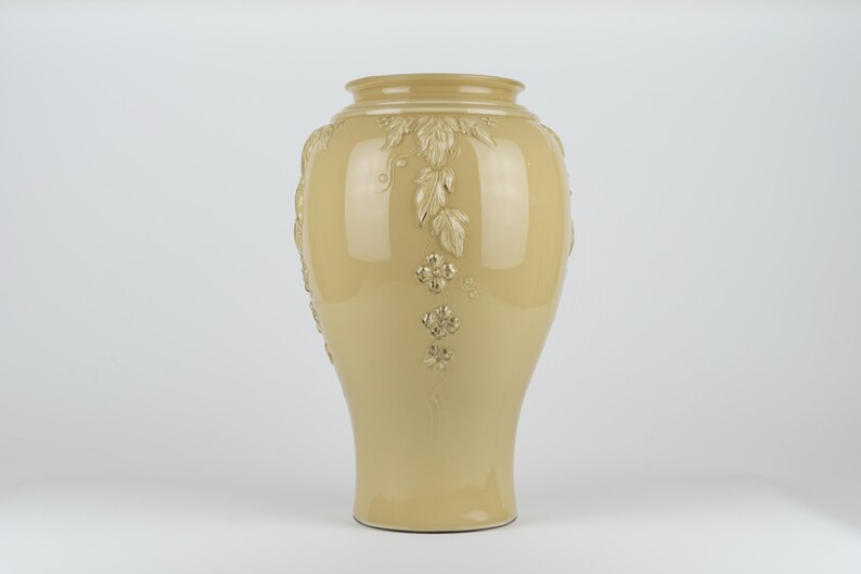Op de afbeelding: Een glanzende, lichtbeige keramische vaas met een brede, ronde buik en een smalle hals. De buitenkant is voorzien van verhoogde, gegoten reli&euml;fdetails van trapsgewijze bladeren en kleine bloemen met vier bloemblaadjes.