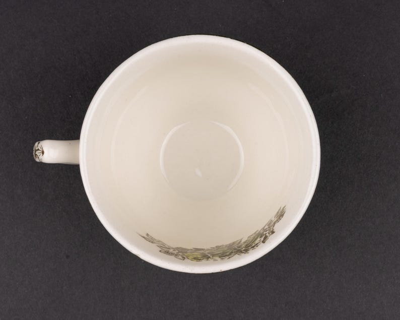Pu&ograve; includere: Una vista dall'alto di una tazza da t&egrave; in ceramica color crema con un manico sul lato sinistro. L'interno della tazza presenta una dettagliata illustrazione botanica verde scuro di fogliame e piccoli fiori lungo la curva inferiore.