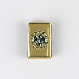 May include: A rectangular gold-toned metal pin featuring an embossed dark blue eagle emblem centered within a circular arrangement of thirteen stars. The design is polished and smooth, with a raised border framing the central patriotic graphic.