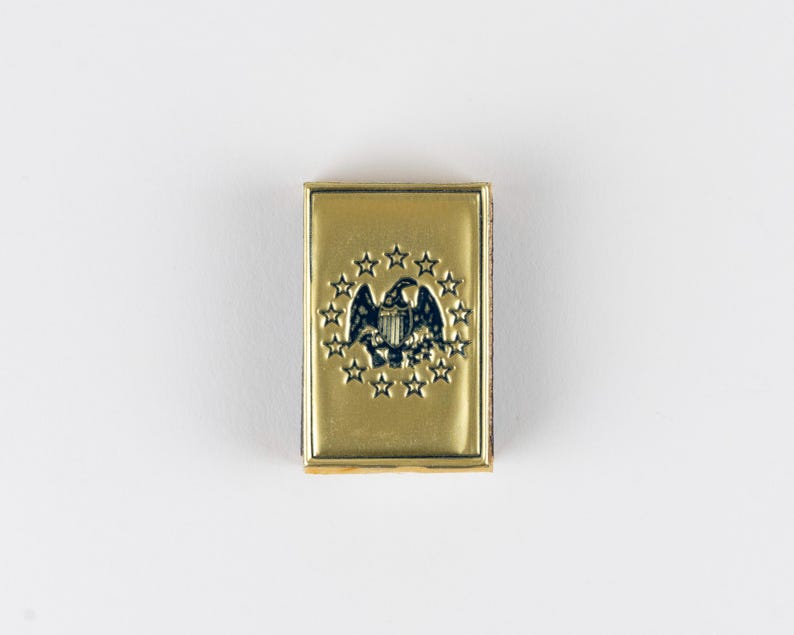 May include: A rectangular gold-toned metal pin featuring an embossed dark blue eagle emblem centered within a circular arrangement of thirteen stars. The design is polished and smooth, with a raised border framing the central patriotic graphic.