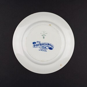 May include: A white ceramic plate with a blue backstamp that reads No 11 Ye Olde Historical Pottery Staffordshire England Rd No 201268. The mark also features an anchor symbol and the letter B. The plate has a smooth, circular rim and a central base.
