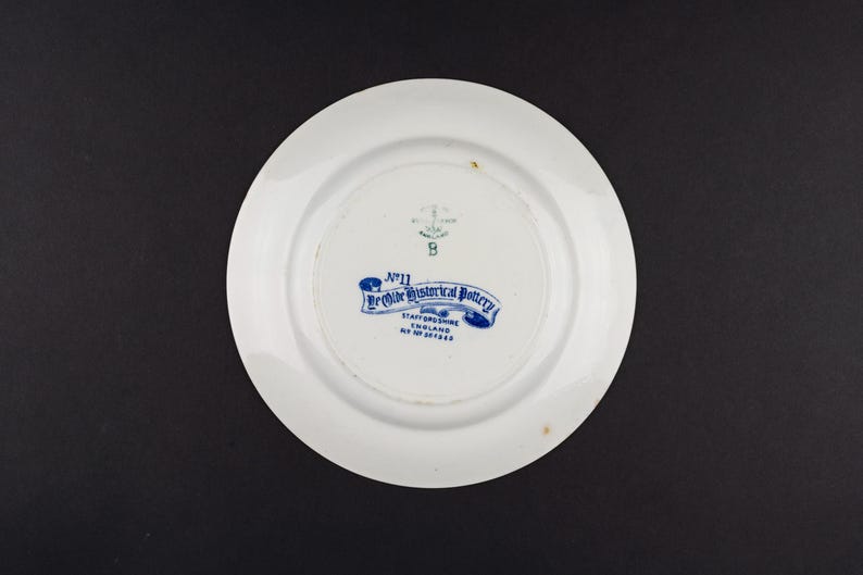 May include: A white ceramic plate with a blue backstamp that reads No 11 Ye Olde Historical Pottery Staffordshire England Rd No 201268. The mark also features an anchor symbol and the letter B. The plate has a smooth, circular rim and a central base.