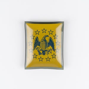 May include: A square glass trinket dish with a yellow center and a dark gray border. The center features a dark blue graphic of an eagle with a shield, arrows, and an olive branch, surrounded by a circular arrangement of thirteen five-pointed stars.