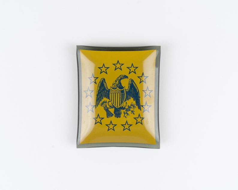 May include: A square glass trinket dish with a yellow center and a dark gray border. The center features a dark blue graphic of an eagle with a shield, arrows, and an olive branch, surrounded by a circular arrangement of thirteen five-pointed stars.