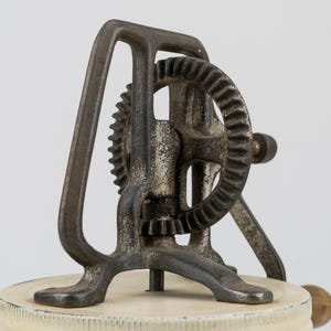 K&ouml;nnte beinhalten: Ein handbetriebenes K&uuml;chenger&auml;t im Vintage-Stil mit einem dunklen, verwitterten Gusseisen-Getriebemechanismus, der auf einem kreisf&ouml;rmigen, cremewei&szlig;en Deckel montiert ist. Das Ger&auml;t verf&uuml;gt &uuml;ber einen vertikalen Rahmen, ein gro&szlig;es Zahnrad und eine seitliche Kurbel.