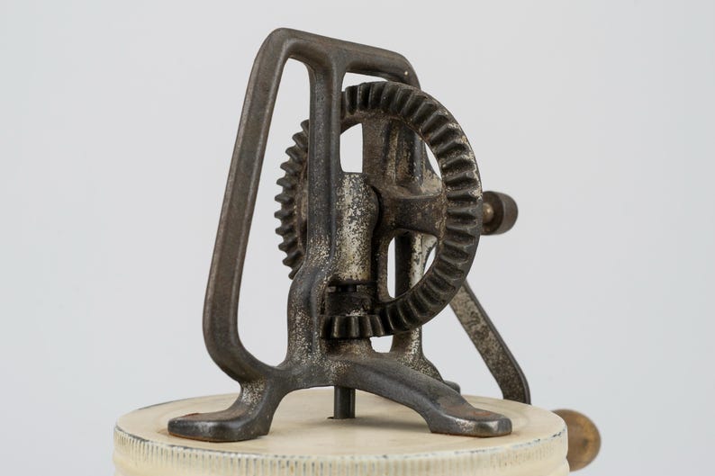 K&ouml;nnte beinhalten: Ein handbetriebenes K&uuml;chenger&auml;t im Vintage-Stil mit einem dunklen, verwitterten Gusseisen-Getriebemechanismus, der auf einem kreisf&ouml;rmigen, cremewei&szlig;en Deckel montiert ist. Das Ger&auml;t verf&uuml;gt &uuml;ber einen vertikalen Rahmen, ein gro&szlig;es Zahnrad und eine seitliche Kurbel.