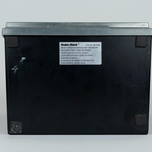 May include: A rectangular black electronic device with a grey top edge and four small square feet on the base. A white label on the back displays the Radio Shack logo, catalog number 60-2420, and instructions requiring two C size batteries.