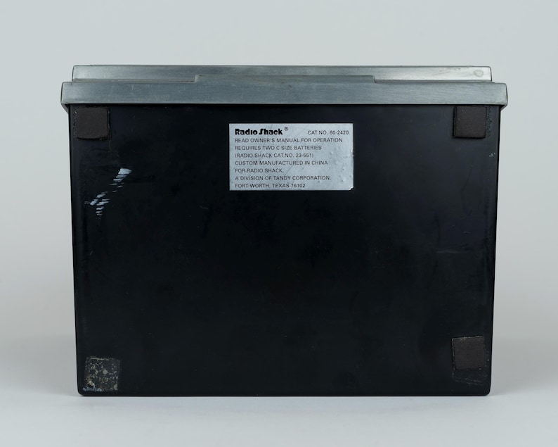 May include: A rectangular black electronic device with a grey top edge and four small square feet on the base. A white label on the back displays the Radio Shack logo, catalog number 60-2420, and instructions requiring two C size batteries.