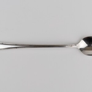 May include: A silver-toned metal spoon featuring an elongated handle with a delicate floral engraving at the tip. The spoon has a smooth, polished bowl and a slender stem that connects to the decorative handle, showcasing a metallic finish.