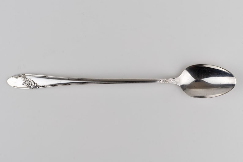 May include: A silver-toned metal spoon featuring an elongated handle with a delicate floral engraving at the tip. The spoon has a smooth, polished bowl and a slender stem that connects to the decorative handle, showcasing a metallic finish.
